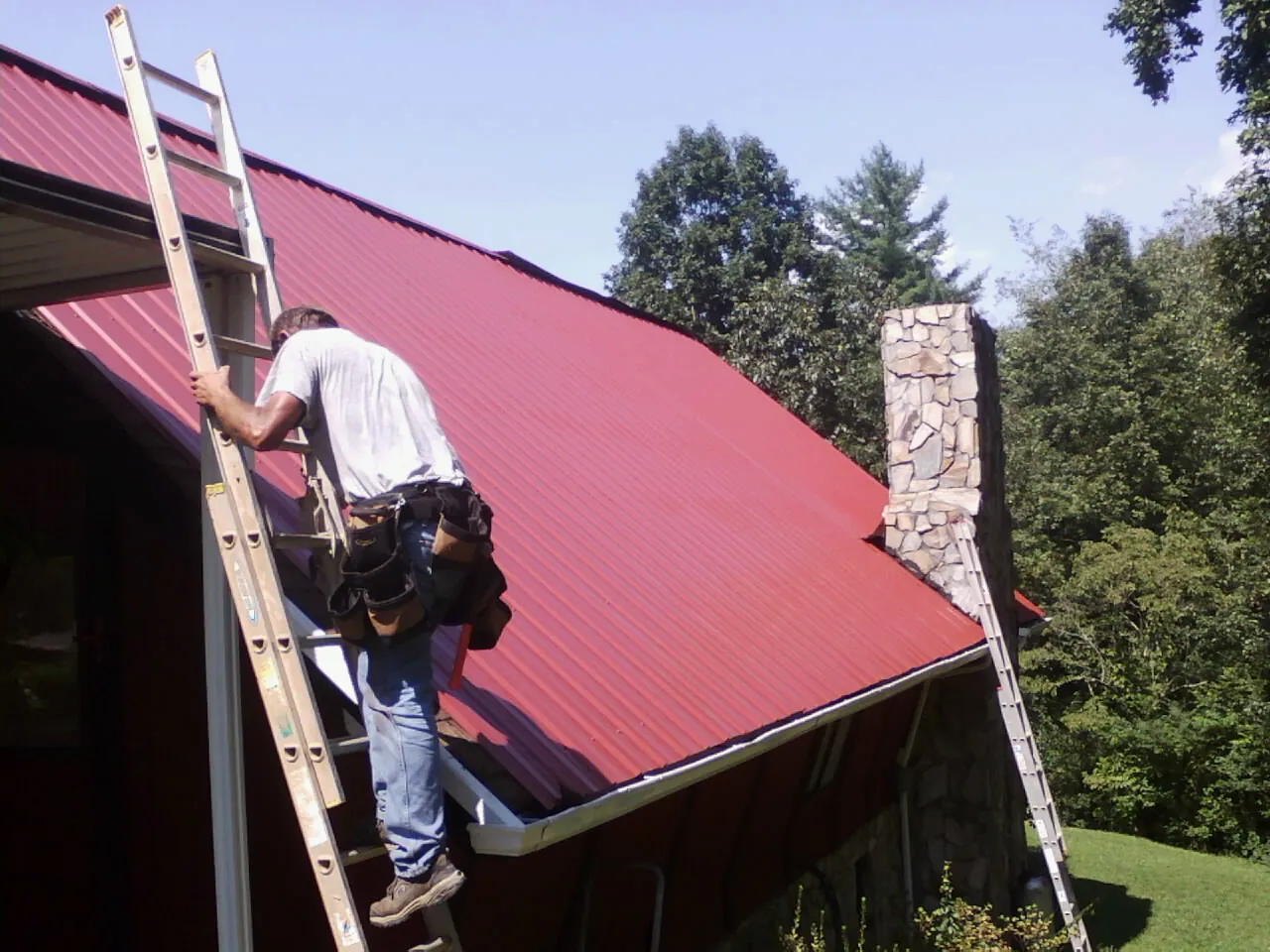 Roofer on a ladder beside a red metal roof and stone chimney — Soffit & Fascia Repair by Roofing Brothers in Bumpus Mills